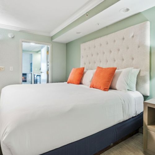 A modern bedroom with a large bed, orange pillows, a stylish lamp, and light-colored decor, creating a cozy and inviting atmosphere.