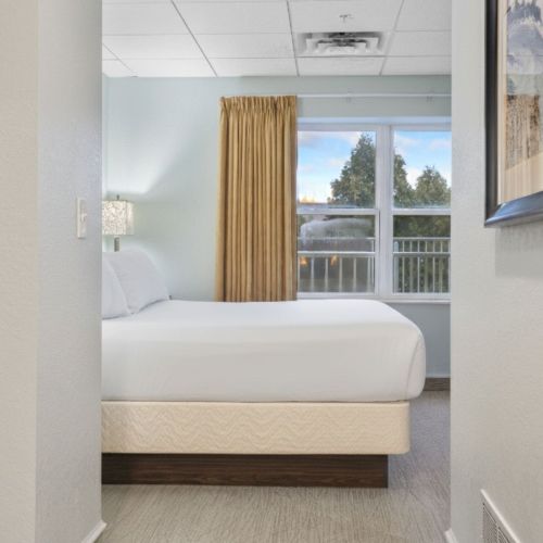 The image shows a minimal hotel room with a bed, beige curtains, window view, neutral walls, and framed artwork on the right side.