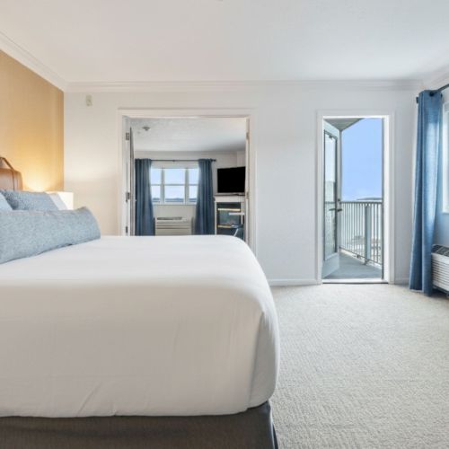 A bright bedroom with a large bed, blue curtains, a dresser, and doors opening to a balcony, creating a cozy and airy atmosphere.