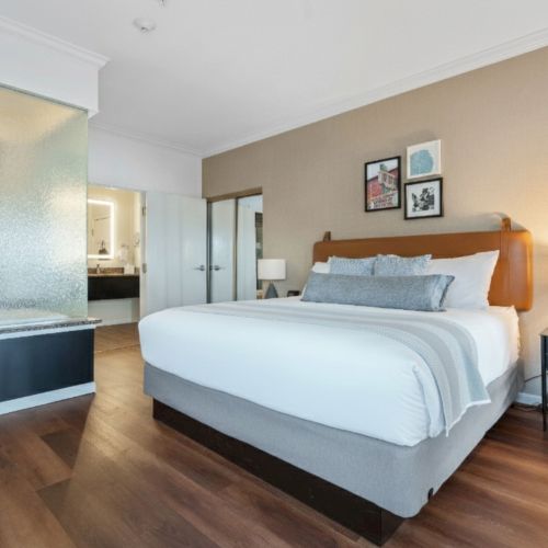 A modern bedroom with a large bed, bedside tables, a semi-open bathroom, and framed wall art on a beige wall.