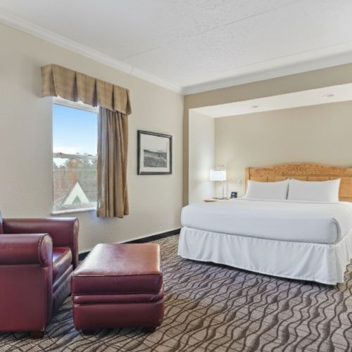 A cozy hotel room with a large bed, two windows, armchair, ottoman, and bedside tables with lamps on a patterned carpet.