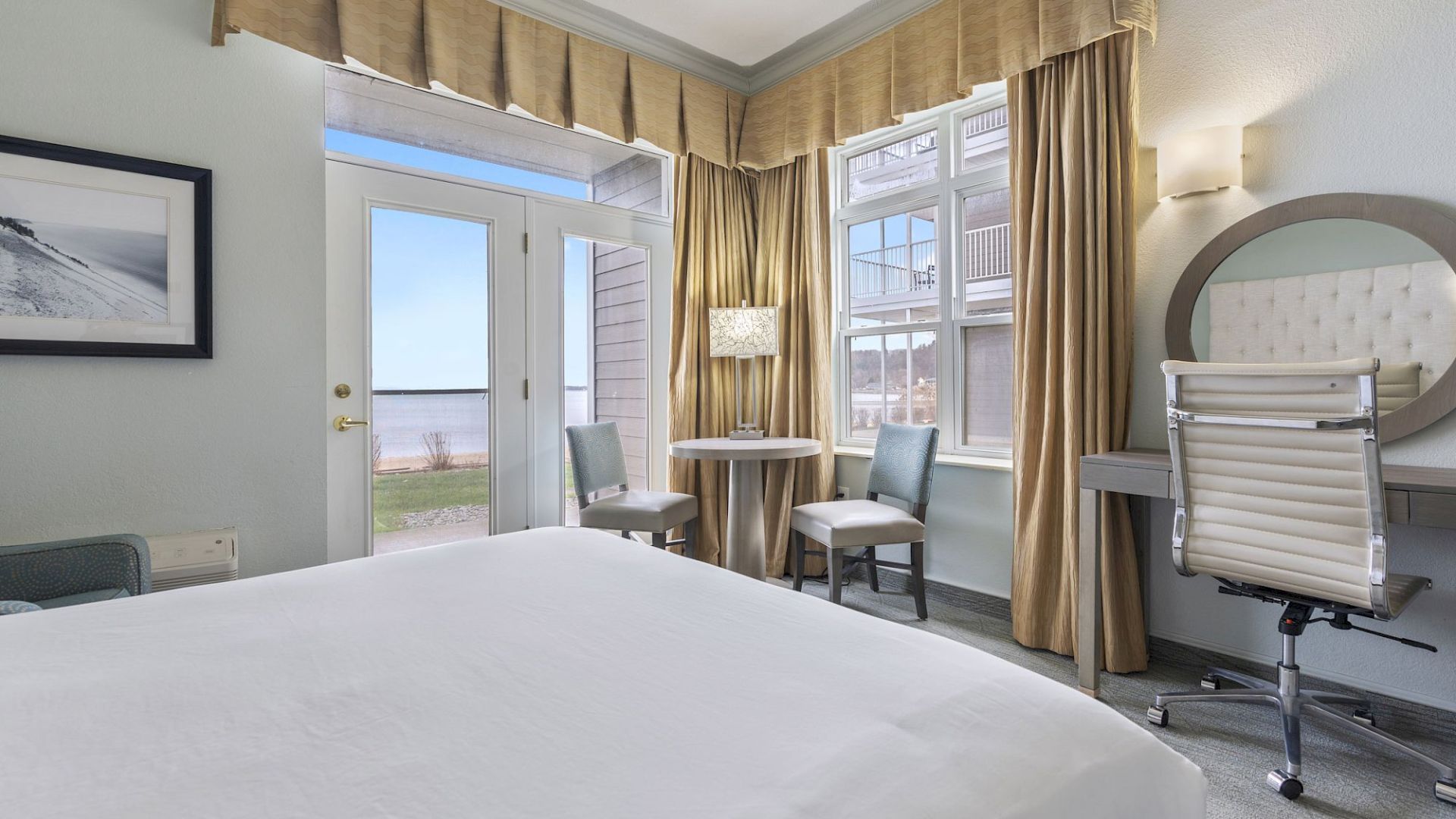 A cozy room with a large bed, desk with chair, small table with two chairs, and a window and door leading outside, overlooking a scenic view.