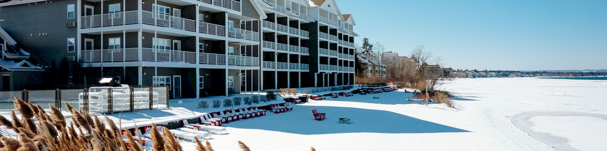 A row of multi-story beachfront buildings with balconies overlooks a snowy landscape and a frozen lake under a clear blue sky.