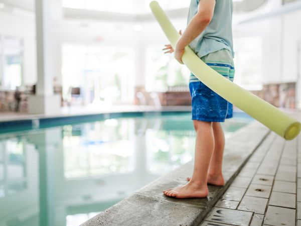A child in swim shorts stands at the edge of an indoor pool, holding a yellow pool noodle, preparing to enter the water.