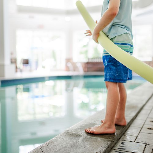 A child stands at the edge of an indoor pool holding a yellow pool noodle, prepared for swimming.