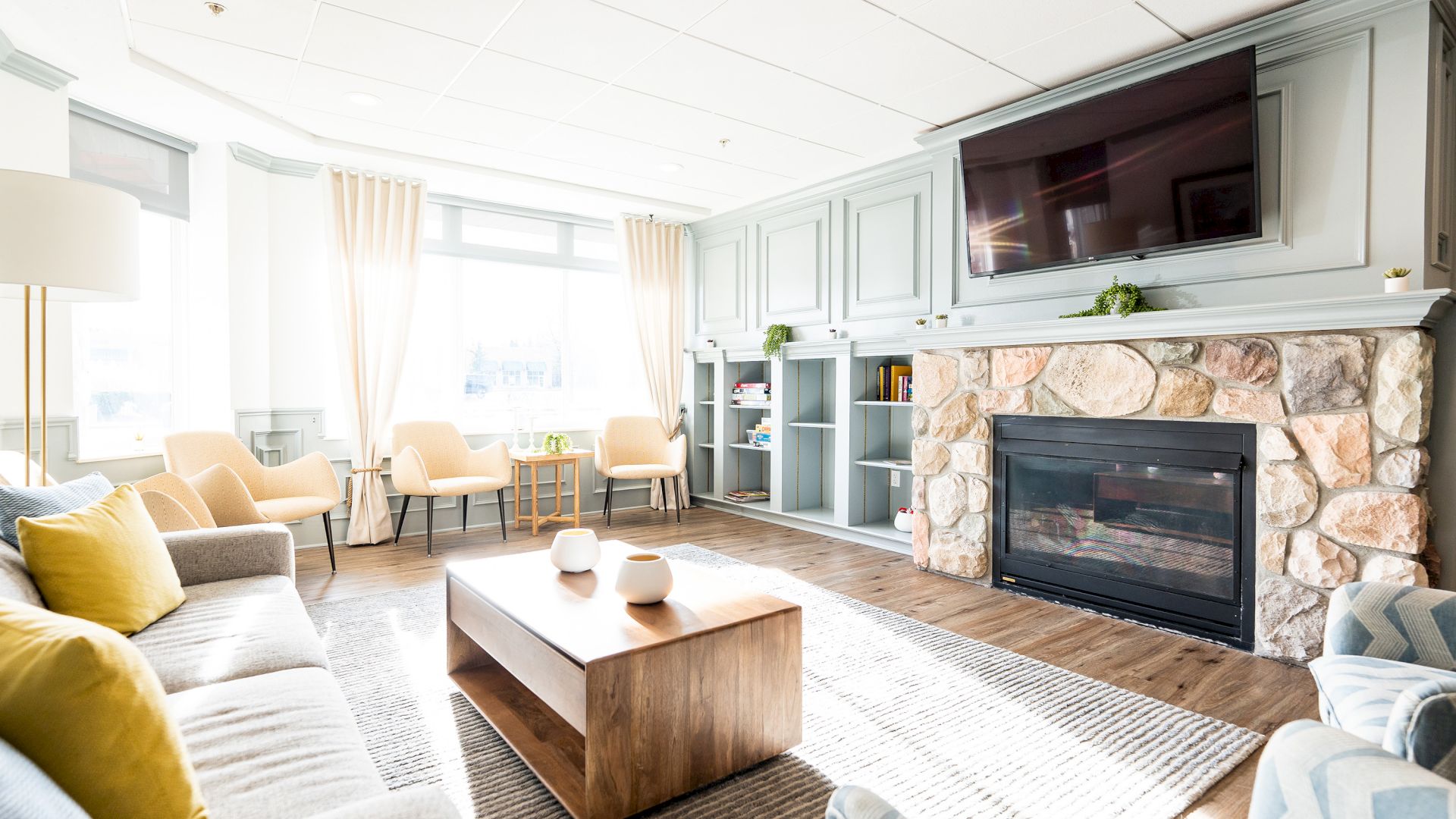 A bright living room with a fireplace, TV, sofas, chairs, a wooden coffee table, and curtains. Shelves and decor create a cozy atmosphere.