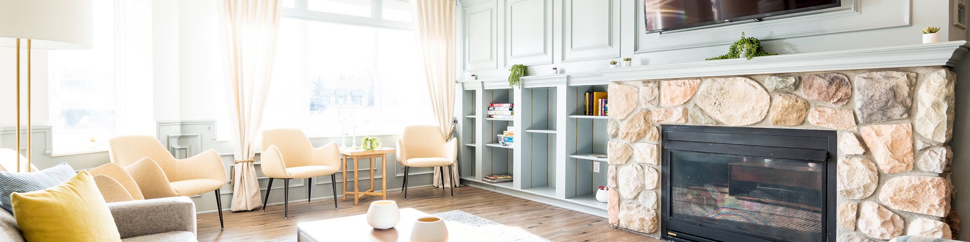 A bright living room with a fireplace, TV, sofas, chairs, a wooden coffee table, and curtains. Shelves and decor create a cozy atmosphere.