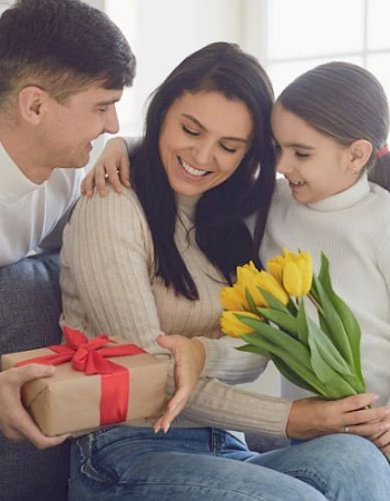 A family sitting on a couch gives a woman a gift wrapped with a red ribbon and a bouquet of yellow tulips in a bright room.