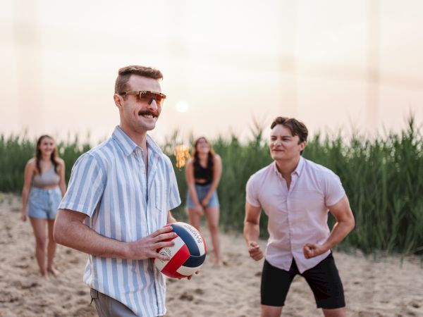 Friends playing beach volleyball at sunset; man with striped shirt holds ball, others ready in the background, tall grasses behind.