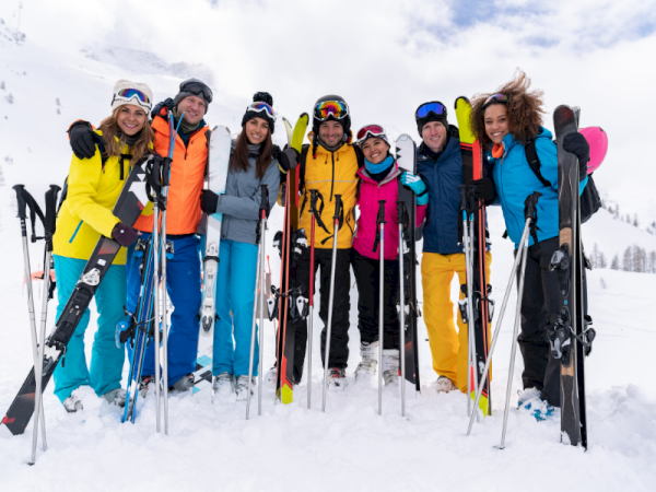 A group of people in colorful ski gear stands on snowy terrain, holding skis and ski poles, smiling for the camera.