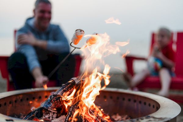 A campfire scene with someone roasting marshmallows on skewers; people in the background are seated in red chairs.