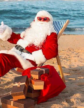 Santa in sunglasses relaxes on a beach chair, surrounded by gifts, giving a thumbs up near the ocean.