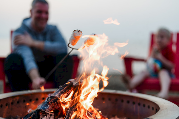 People are sitting by a campfire, roasting marshmallows on skewers, with a relaxed vibe.