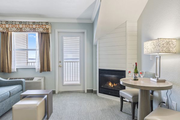 A cozy room with a fireplace, small table, two chairs, sofa, and a windowed door leading outside. Two wine glasses and a bottle are on the table.