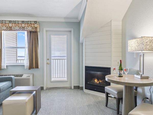A cozy room with a fireplace, small table, two chairs, sofa, and a windowed door leading outside. Two wine glasses and a bottle are on the table.