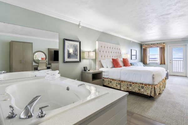 A hotel room features a large bed, a whirlpool tub, stylish decor, and a view through glass doors leading to a balcony area.