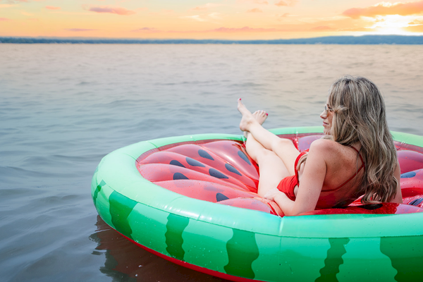 A person relaxes on a watermelon-themed float on a calm body of water during sunset, wearing sunglasses and enjoying the view.