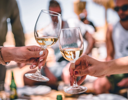 Two people clink wine glasses in a social, outdoor gathering scene, surrounded by casually dressed individuals.