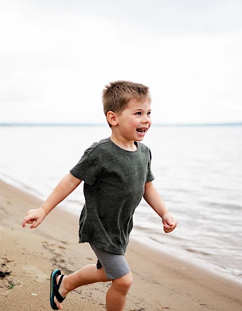 A joyful boy runs along a sandy beach by the water, wearing a dark shirt, shorts, and sandals, with a big smile as waves lap the shore.