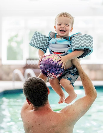 A father tosses his joyful baby into the air over a swimming pool, both smiling as sunlight fills the indoor pool area.