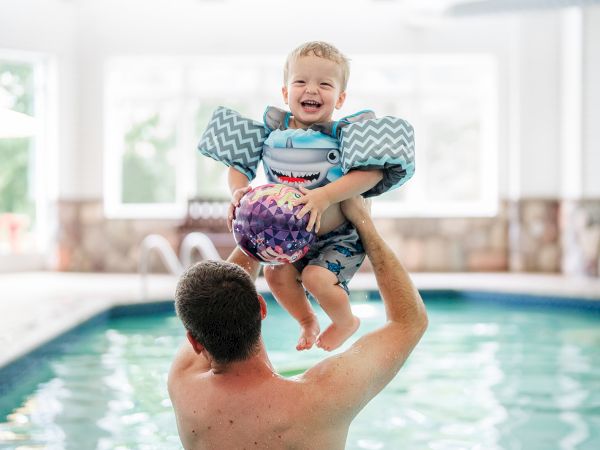 A child in swim gear is joyfully lifted into the air by an adult at an indoor pool, both smiling as water sparkles nearby.