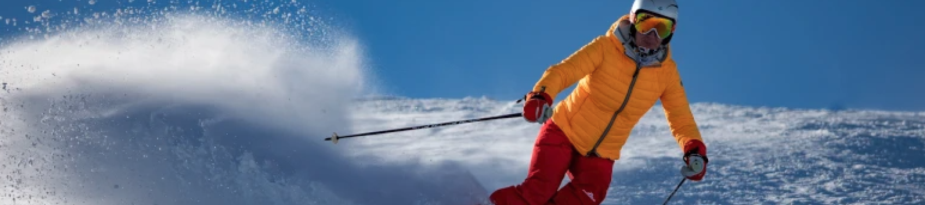 A skier in a yellow jacket and red pants glides down a snowy slope, kicking up powder under a clear blue sky.