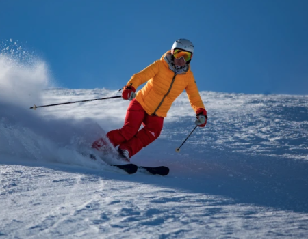 A skier in a yellow jacket and red pants glides down a snowy slope, kicking up powder under a clear blue sky.