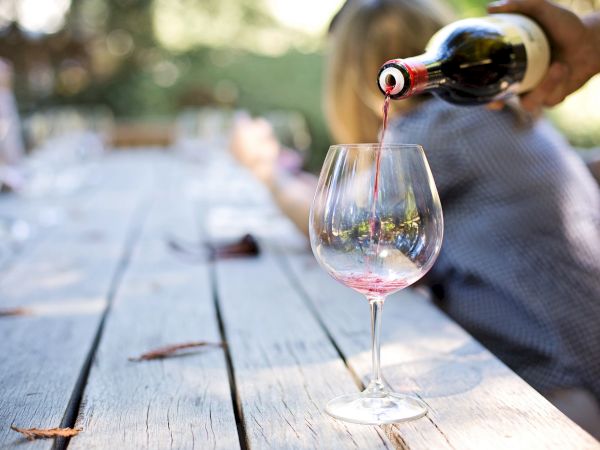 A hand pours red wine into a glass on a wooden outdoor table; the background features people in a relaxed, blurred setting.
