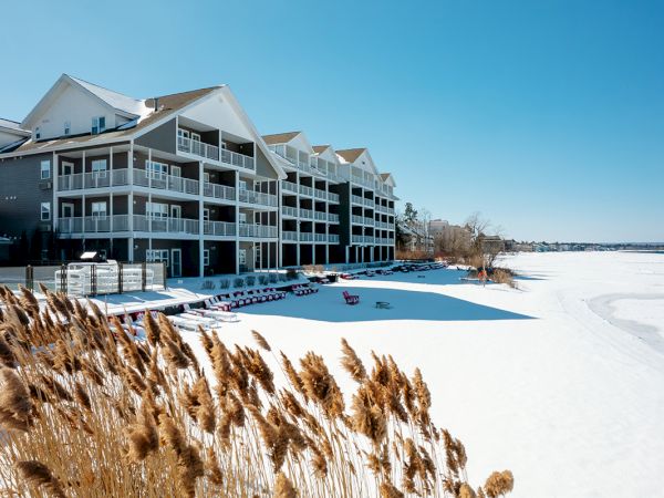 Snowy beach with a multi-story hotel, a frozen shoreline, and tall grasses in the foreground; clear blue sky overhead.