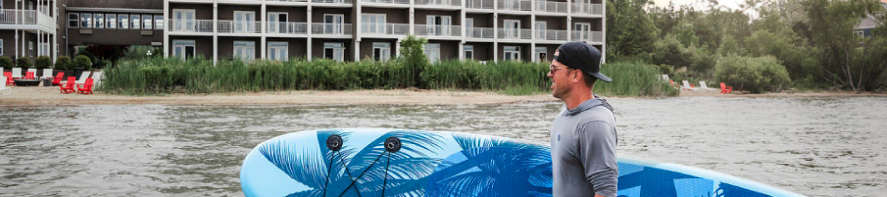 A man stands in ankle-deep water holding a blue windsurf/ SUP board near a lake with a multi-story hotel building and sunset behind.