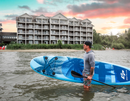 A man stands in ankle-deep water holding a blue windsurf/ SUP board near a lake with a multi-story hotel building and sunset behind.