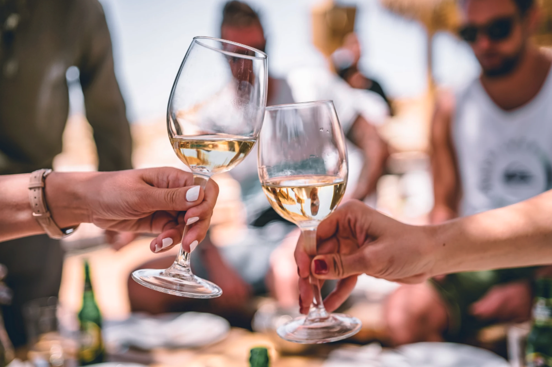 People cheers with wine glasses at a sunny outdoor gathering, hands clinking over a table of drinks and snacks.