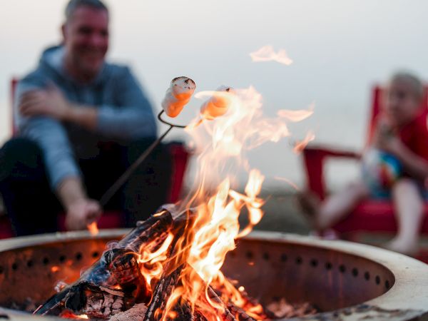 A campfire with glowing embers, roasting marshmallows on sticks, and people in the background near a beach setting.