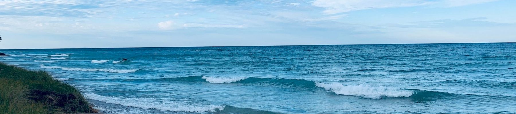 A tranquil shore with gentle waves, turquoise sea, blue sky, wispy clouds, and green dune grass on the left by a sandy beach.