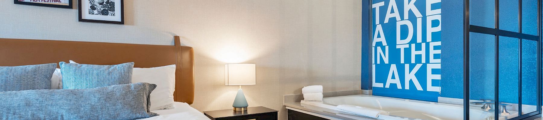 A modern hotel room with a large bed, nightstand, lamp, and framed art; a blue accent wall with a bold &ldquo;TAKE A DIP IN THE LAKE&rdquo; sign by a glass-enclosed tub.