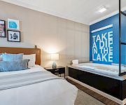 A modern hotel room with a large bed, nightstand, lamp, and framed art; a blue accent wall with a bold “TAKE A DIP IN THE LAKE” sign by a glass-enclosed tub.