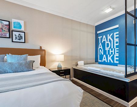 A modern hotel room with a large bed, nightstand, lamp, and framed art; a blue accent wall with a bold &ldquo;TAKE A DIP IN THE LAKE&rdquo; sign by a glass-enclosed tub.