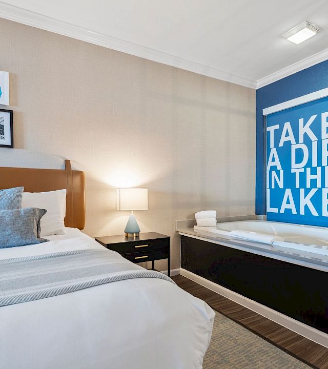 A modern hotel room with a large bed, nightstand, lamp, and framed art; a blue accent wall with a bold “TAKE A DIP IN THE LAKE” sign by a glass-enclosed tub.