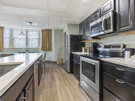 A modern kitchen with dark cabinets, stainless steel appliances (fridge, oven, microwave), a long island, and light wood flooring.