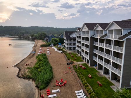 Aerial view of a lakeside resort: multi-story beige buildings along the shore, sandy beach with lounge chairs, and calm water at sunset.