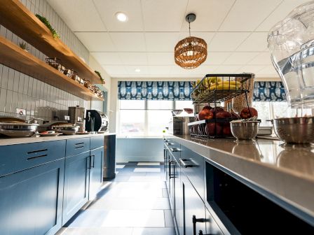 Cozy, bright cafe kitchen with blue cabinets, white tile, hanging basket light, open shelves, and a sunny window at the far end.