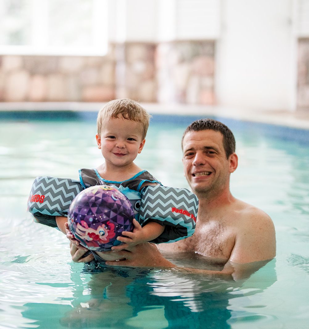 A man and a child are swimming in a pool, smiling as they hold a purple ball together.