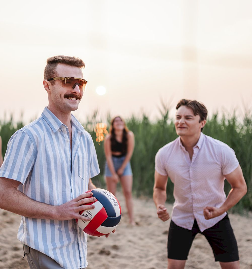 A group of friends playing beach volleyball on a sunny day; two players near the net ready to serve, others in the background by tall grass.