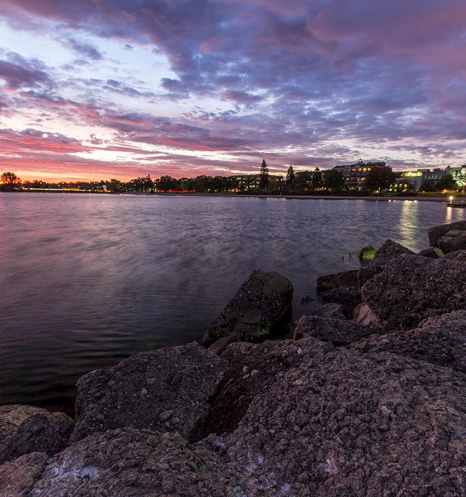 A rocky shoreline by a calm lake at sunset; purple-pink clouds glow over distant buildings, water reflecting the colorful sky.