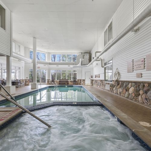 A spacious indoor pool area with palm-like seating, a hot tub in the foreground, large windows, white walls, and a stone-accent wall along the edge.