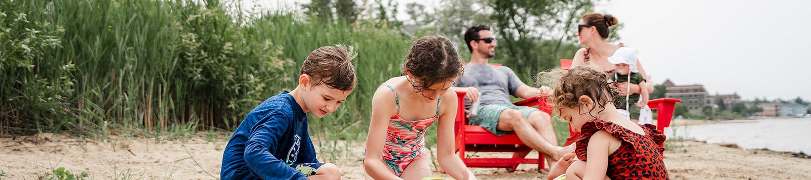 Kids play on a sandy beach with buckets and shovels, while adults relax on chairs nearby and a building sits beyond the tall grass.