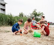 Kids play on a sandy beach with buckets and shovels, while adults relax on chairs nearby and a building sits beyond the tall grass.