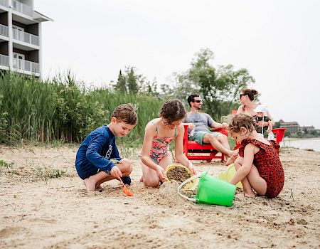 Kids play on a sandy beach with buckets and shovels, while adults relax on chairs nearby and a building sits beyond the tall grass.