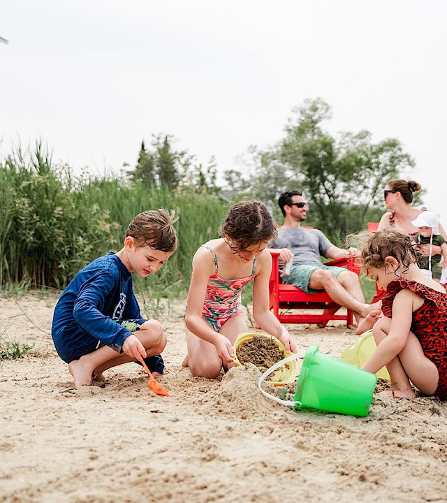 Kids play on a sandy beach with buckets and shovels, while adults relax on chairs nearby and a building sits beyond the tall grass.
