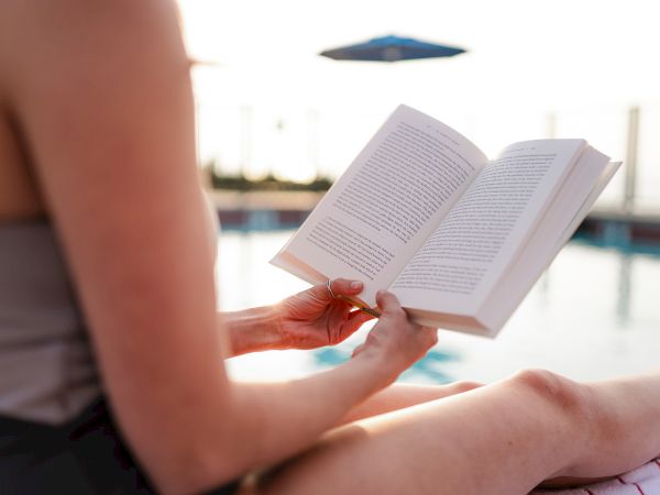 A person swimming poolside, sitting and reading a paperback book with a dotted line of pages visible, while a distant umbrella and water backdrop gleam.